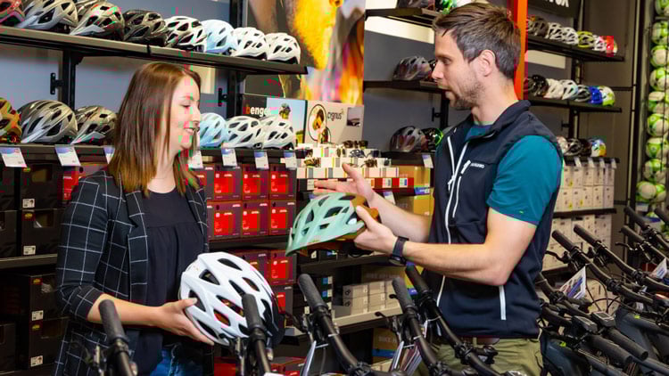 Zwei Personen in einem Fahrradladen betrachten Fahrradhelme. Die Person links hält einen weißen Helm, die Person rechts einen hellblauen. Im Hintergrund sind Regale mit weiteren Helmen und Fahrradzubehör zu sehen