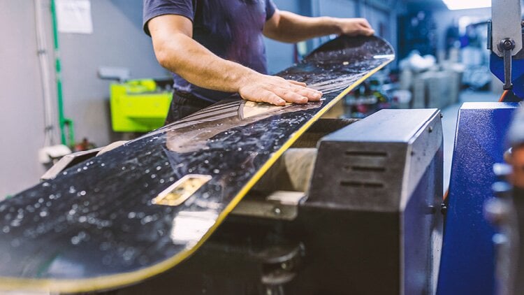 Eine Person arbeitet an einem Snowboard. Die Person verwendet eine Maschine, um die Unterseite des Snowboards zu reparieren. Die Hände der Person liegen auf dem Snowboard und führen es durch die Maschine. Im Hintergrund sind verschiedene Werkzeuge und Ausrüstungen zu sehen, die typischerweise in einer Werkstatt oder Reparaturwerkstatt zu finden sind.