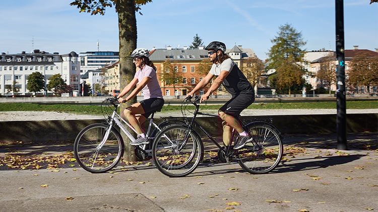 Zinsswitch Zinsswitch. Zwei personen in sportlichen Outfits fahren Fahrrad an einer Alle. Im Hintergrund sind Bäume und Häuser.