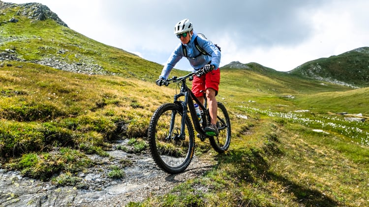 Person auf einem Mountainbike fährt auf einem schmalen, steinigen Pfad durch hügelige, grasbewachsene Landschaft; trägt blauen Langarmshirt, rote Shorts, Helm und Rucksack. Im Hintergrund grüne Hügel und teils bewölkter Himmel.