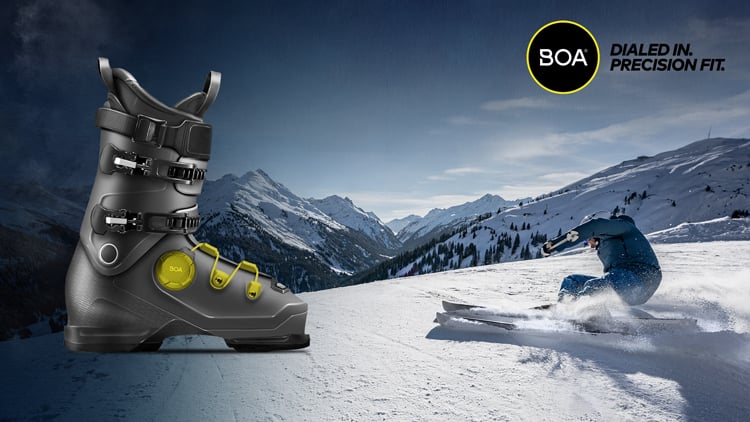 Skischuh mit BOA-Logo im Vordergrund und dem Text ‚DIALED IN. PRECISION FIT.‘ oben rechts. Im Hintergrund fährt ein Skifahrer eine scharfe Kurve auf einem verschneiten Berghang, mit weiteren Bergen in der Ferne.