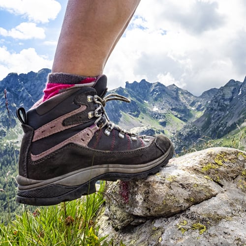 Wanderschuh auf Felsen vor alpiner Bergkulisse mit bewölktem Himmel.