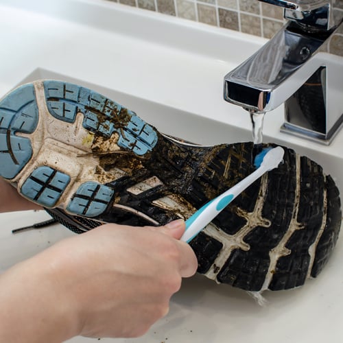 Laufschuhe reinigen Eine Person reinigt die verschmutzte Sohle eines Schuhs mit einer Zahnbürste unter fließendem Wasser aus einem Wasserhahn. Die Sohle ist mit Schlamm und Schmutz bedeckt