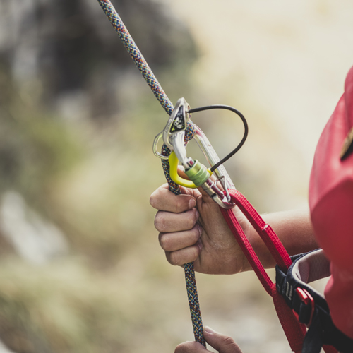 Nahaufnahme einer Person mit Kletterausrüstung, die ein Seil mit einer Hand hält. Das Seil ist durch ein Sicherungsgerät geführt, das am Klettergurt befestigt ist. Karabiner sind ebenfalls an der Ausrüstung zu sehen.