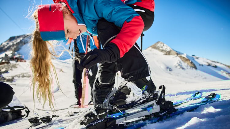 Eine Person steht auf verschneitem Boden in den Bergen und stellt die Bindung ihrer Skier ein. Sie trägt eine blau-rote Jacke mit roten Ärmeln, schwarze Handschuhe, schwarze Hose, Skischuhe sowie ein rot-blaues Stirnband. Im Hintergrund sind weitere Skifahrer und ein klarer blauer Himmel zu sehen.