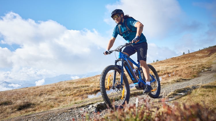 Person auf einem blauen E-Mountainbike fährt auf einem Trail in bergiger Landschaft; trägt Helm, Sonnenbrille und Outdoor-Radbekleidung. Im Hintergrund eine malerische Aussicht mit Wolken und entfernten Bergen.