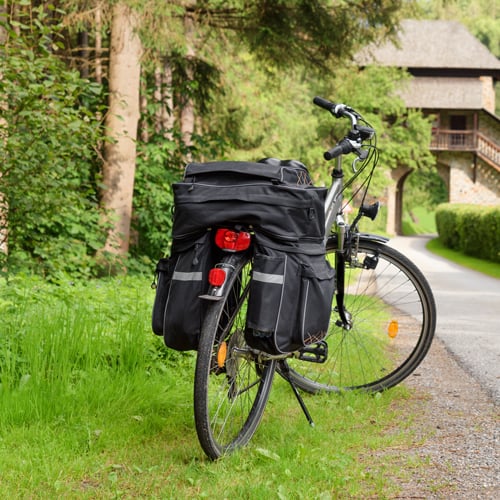 Fahrrad mit mehreren schwarzen Gepäcktaschen am hinteren Gepäckträger steht am Rand eines asphaltierten Wegs in grüner Umgebung; im Hintergrund ein rustikales Gebäude mit Steinmauern und Holzelementen, teils von Bäumen verdeckt.