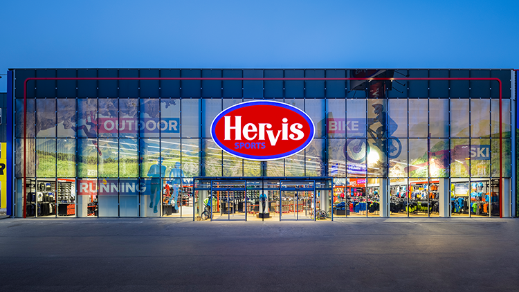 Die Außenansicht des Hervis Sports-Stores in Wien Stadlau. Die Fassade besteht aus Glaswänden, und in der Mitte befindet sich ein großes rotes und blaues Hervis Sports-Logo. Über dem Eingang sind die Wörter 'OUTDOOR', 'BIKE', 'SKI' und 'RUNNING' auf den Glaswänden zu sehen. Durch das Glas ist das Innere des Geschäfts mit verschiedenen Sportgeräten und -bekleidung sichtbar.