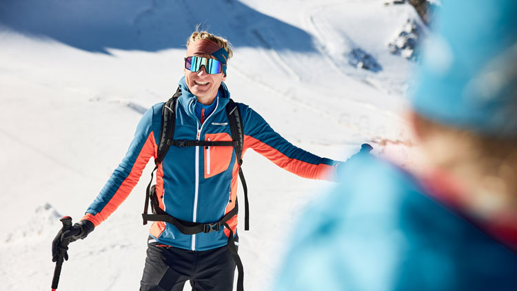 Eine Person in blauer und orangefarbener Winter-Sportkleidung steht mit Skistöcken auf einem verschneiten Hang. Im Vordergrund ist eine weitere Person in ähnlicher Kleidung teilweise sichtbar