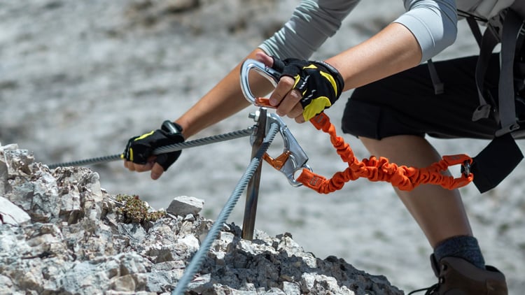 Eine Person trägt Handschuhe und Outdoor-Ausrüstung und sichert einen Karabiner an einem Sicherheitsseil auf einer felsigen Oberfläche.