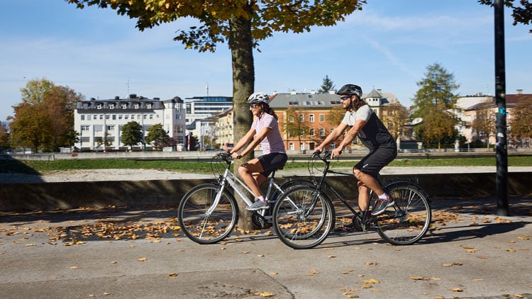 Zwei Personen fahren mit Citybikes auf einem asphaltierten Weg in parkähnlicher Umgebung; im Hintergrund herbstlich gefärbte Bäume, Gebäude und blauer Himmel. Beide tragen Helme und Fahrradkleidung der Marke Benger.