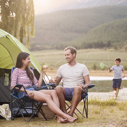 Zwei Personen sitzen in Campingstühlen vor einem grünen Zelt und halten Getränke in der Hand. Im Hintergrund läuft ein Kind mit einem Kescher in der Nähe eines Gewässers. Dahinter sind Bäume und Berge zu sehen.