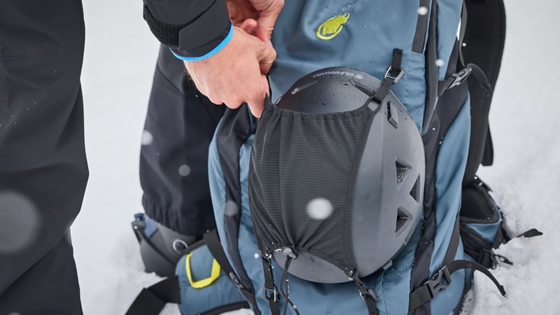 Eine Person befestigt einen schwarzen Skihelm außen an einem blauen Skitourenrucksack