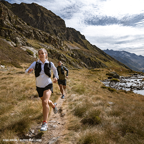 Zwei Trailrunner auf schmalem Pfad durch bergige Graslandschaft, umgeben von Felsen und Bergen unter teils bewölktem Himmel; beide tragen Sportkleidung und kleine Rucksäcke. Foto: ©SALOMON / ALEXIS BERG