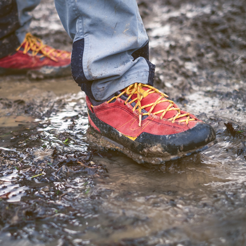 Eine Person steht mit rot-gelben Wanderschuhen in einem schlammigen, nassen Gelände. Die hellblauen Hosenbeine sind leicht hochgekrempelt, und die Schuhe sind teilweise mit Matsch bedeckt.