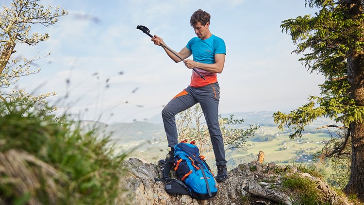 Person steht auf einem felsigen Vorsprung in der Natur, vermutlich beim Wandern oder Klettern. Sie trägt Outdoor-Kleidung mit blauem Shirt und grauer Hose mit orangen Akzenten, hält ein Wanderstock in der Hand. Neben ihr liegt ein großer blauer Rucksack. Im Hintergrund sind Bäume sowie eine hügelige Landschaft unter teils bewölktem Himmel zu sehen