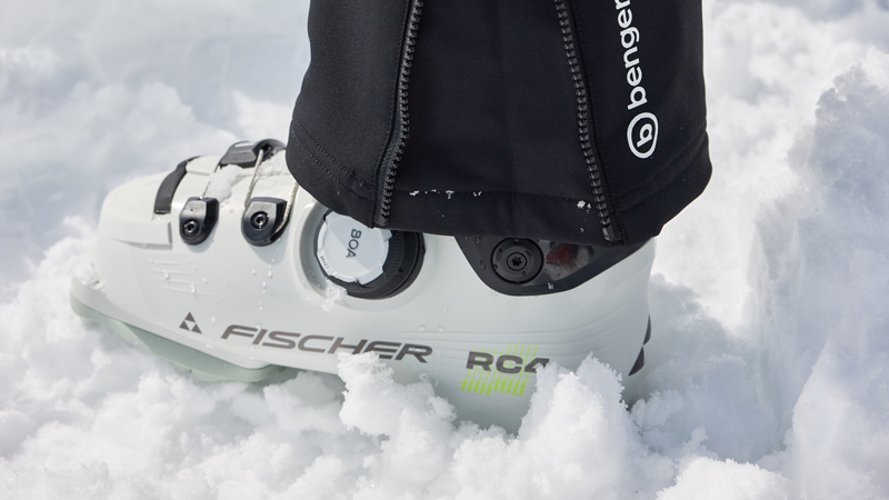 Skischuhe richtig pflegen Eine Person trägt einen weißen Fischer RC4-Skischuh mit BOA-System und drei Schnallen im Schnee, das schwarze Hosenbein der Marke Benger ist gut sichtbar.