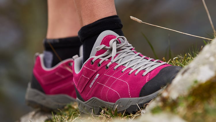 Nahaufnahme von zwei Füßen in pink-grauen Wanderschuhen der Marke Kilimanjaro, getragen mit schwarzen Socken. Die Schuhe stehen auf einem gemischten Untergrund aus Gras und Steinen