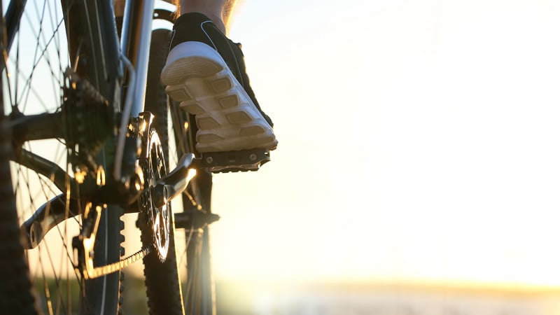 Nahaufnahme eines Fußes in einem schwarz-weißen Sportschuh auf dem Pedal eines Fahrrads. Die Szene ist von Sonnenlicht durchflutet.