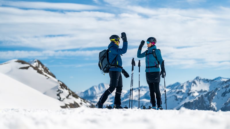 Zwei Skitourengeher geben sich auf einem verschneiten Berggipfel ein High Five.