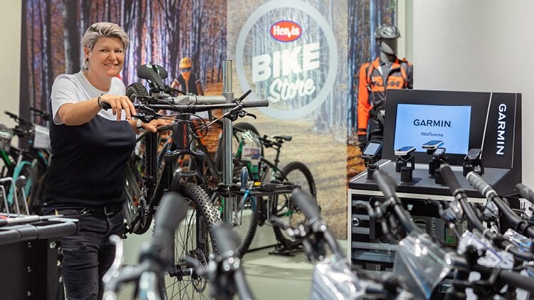 Das Innere eines Hervis BIKE Stores. Mehrere Fahrräder sind ausgestellt, und eine Person hält eines der Fahrräder. Im Hintergrund steht eine Schaufensterpuppe, die in Fahrradbekleidung gekleidet ist. Der Hintergrund zeigt ein Bild eines Waldes mit Radfahrern, die hindurchfahren.