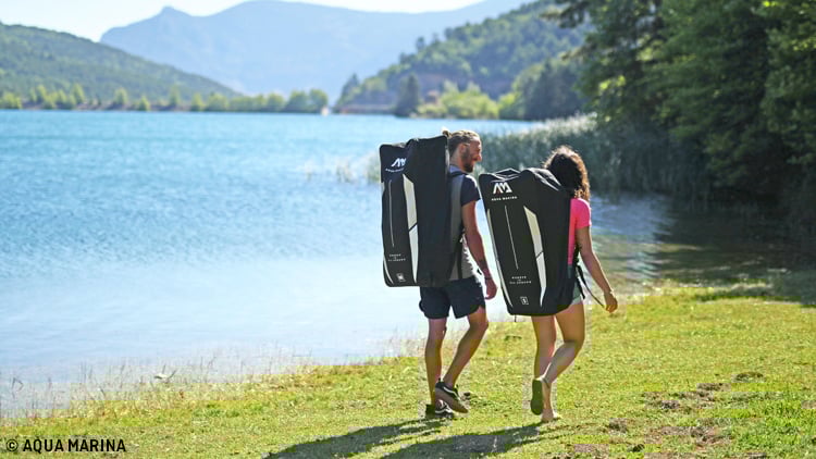 Zwei Personen gehen am Ufer eines Sees entlang und tragen große schwarze Aqua Marina-Rucksäcke. Im Hintergrund sind ein ruhiger See, bewaldete Ufer und Berge zu sehen.