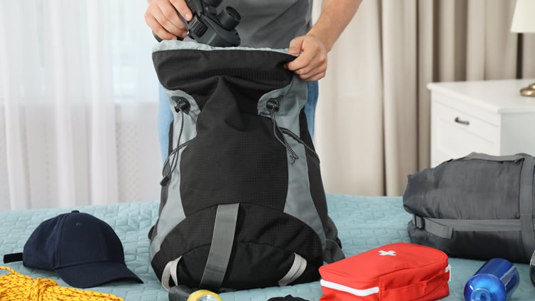 Person packt einen Rucksack auf einem Bett, umgeben von Outdoor-Ausrüstung wie Kappe, Seil, Taschenlampe und Erste-Hilfe-Set.