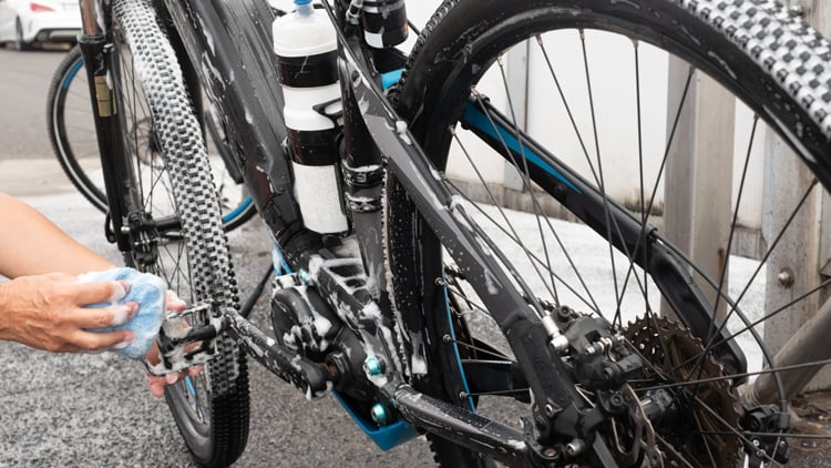 Person reinigt ein E-Bike im Außenbereich mit einem blauen Schwamm; das Rad ist mit Seifenschaum bedeckt. Im Hintergrund sind Pflastersteine und Gebäude zu sehen.
