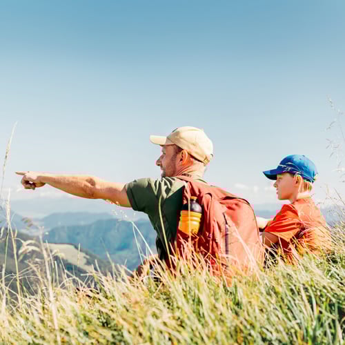 Zwei Personen mit Rucksäcken sitzen im Gras und blicken auf eine Berglandschaft, eine zeigt in die Ferne.