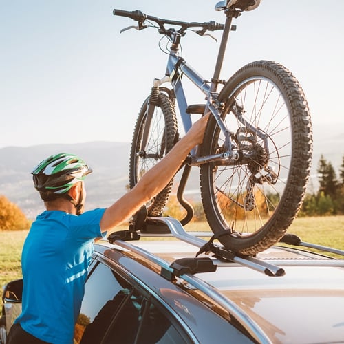 Person mit Helm und blauem Shirt befestigt ein Fahrrad auf einem Dachträger eines Autos; im Hintergrund Bäume und hügelige Landschaft.