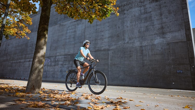 E-Bike Kaufberatung Person mit Helm, hellblauem Shirt und schwarzen Shorts fährt auf einem mit Herbstlaub bedeckten Asphaltweg mit einem E-Bike; links ein Baum mit herbstlich gefärbten Blättern, im Hintergrund eine große Betonwand und blauer Himmel.