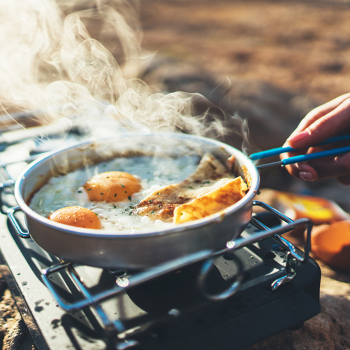 Nahaufnahme einer Bratpfanne auf einem tragbaren Campingkocher mit zwei Eiern und weiteren Speisen. Dampf steigt auf. Der Hintergrund deutet auf eine Outdoor-Umgebung hin.