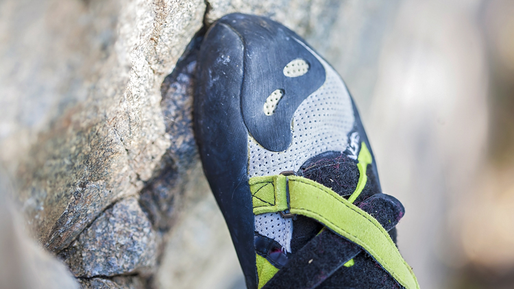 Nahaufnahme eines Kletterschuhs, der gegen eine Felsoberfläche gedrückt wird. Der Schuh hat eine schwarze Gummispitze, ein graues Mesh-Obermaterial mit schwarzen und gelben Akzenten sowie grüne Verschlussriemen.