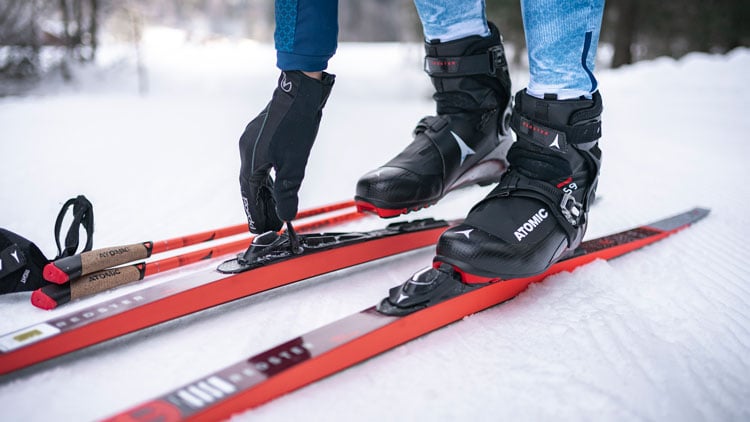 Nahaufnahme einer Person, die schwarze Skischuhe und blaue Hosen trägt und die Bindungen an roten Langlaufskiern mit Handschuhen befestigt. Neben den Skiern liegen zwei Skistöcke mit roten Griffen im Schnee. Die Marke ‚ATOMIC‘ ist auf den Skischuhen und Skiern sichtbar