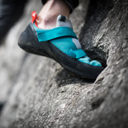 Nahaufnahme eines Fußes in einem türkis-schwarzen Kletterschuh mit roten Akzenten auf einer rauen Felsoberfläche – beim Klettern.