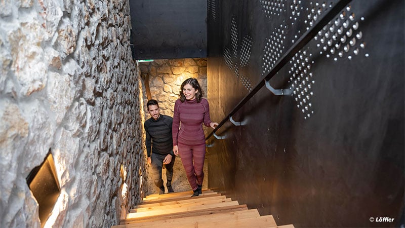 Welche Funktionsunterwäsche ist die beste? | © Löffler Zwei Personen gehen eine Treppe hinauf, die links von einer Steinmauer mit eingebauten Leuchten und rechts von einer dunklen Wand mit dekorativen Mustern begrenzt wird. Beide sind in Freizeitkleidung unterwegs. Foto: Löffler