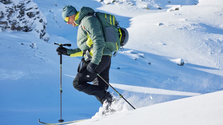 Person steigt mit Skiern und Skistöcken einen verschneiten Berghang hinauf. Sie trägt eine grüne Jacke, schwarze Hose, Handschuhe, Mütze und einen großen Rucksack. Im Hintergrund sind schneebedecktes Gelände und vereinzelte Felsen zu sehen