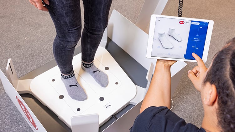 Person steht mit Socken auf einer weißen Fußmessplatte, während eine zweite Person ein Tablet mit einer 3D-Fußscan-Ansicht bedient.