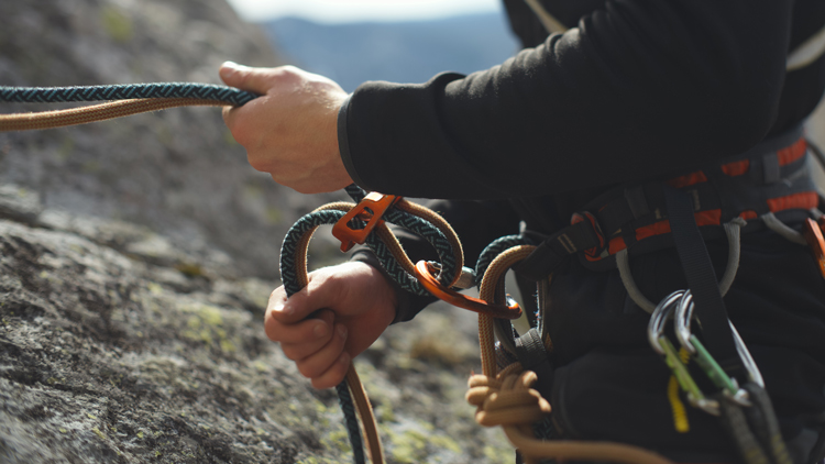 Nahaufnahme einer Person beim Klettern: Sie trägt schwarze Kleidung und verwaltet mit beiden Händen Kletterseile, die mit Karabinern und weiterem Kletterequipment am Gurt befestigt sind. Im Hintergrund ist eine Felswand zu sehen.