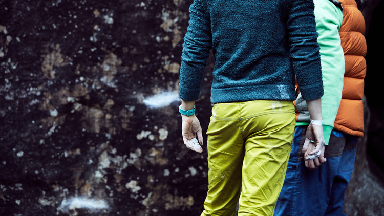 Zwei Personen stehen nebeneinander vor einer Felswand. Eine trägt ein blaues Langarmshirt mit gelber Hose, die andere eine orange Steppweste über einem hellgrünen Shirt und blaue Hose. Beide haben Chalk an den Händen, was auf Kletteraktivitäten hinweist. Die Felswand im Hintergrund zeigt weiße Chalkspuren