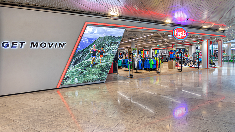 Die Außenansicht des Hervis Stores Europark Salzburg. Der Store hat einen großen Glaseingang mit dem Hervis-Logo, das gut sichtbar darüber angebracht ist. Vor dem Store befindet sich ein Parkplatz mit mehreren Büschen und einem Fußgängerüberweg, der zum Eingang führt. Im Hintergrund sind unter einem klaren blauen Himmel Berge zu sehen.