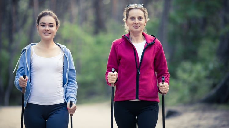 Zwei Personen beim Nordic Walking auf einem von Grün umgebenen Weg. Eine trägt ein weißes Tanktop mit blauer Kapuzenjacke, die andere eine rote Jacke über einem weißen Shirt. Beide halten Walking-Stöcke