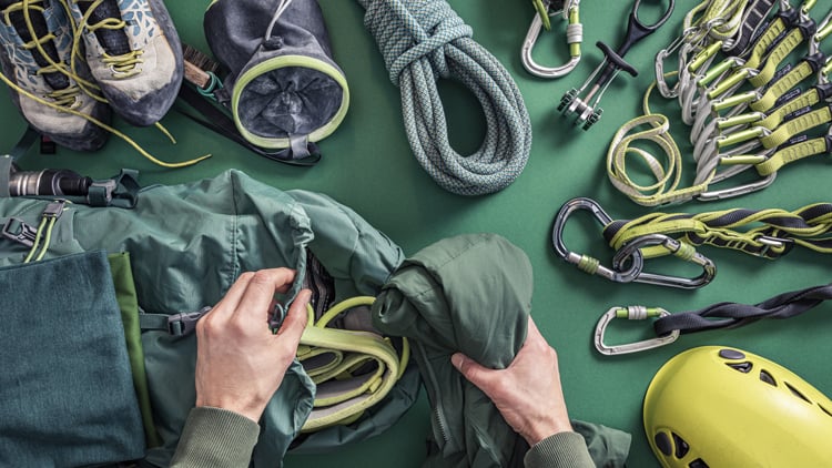 Verschiedene Kletterausrüstungsgegenstände liegen auf einer grünen Fläche: Kletterschuhe, Chalkbag, ein aufgerolltes Seil, Karabiner, Expresssets und ein gelber Helm. 