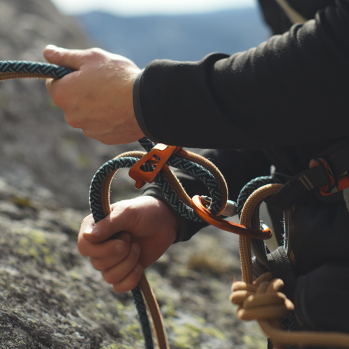 Eine Person mit schwarzem Langarmshirt hält blaue und beige Kletterseile in beiden Händen. Die Seile sind durch ein orangefarbenes Sicherungsgerät geführt. Im Hintergrund sind Felsen und verschwommene Berglandschaft zu erkennen.