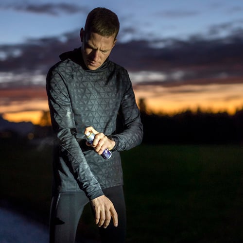 Person in dunklem Kapuzenpullover mit Muster und schwarzer Hose steht im Freien bei Sonnenuntergang und sprüht mit einer Aerosoldose auf den Arm; das Gesicht ist unscharf