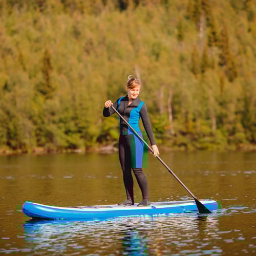 Eine Person steht auf einem blauen Paddleboard im Wasser und hält ein Paddel. Sie trägt einen Neoprenanzug. Im Hintergrund sind grüne Bäume und dichte Vegetation zu sehen