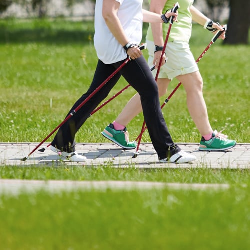 Zwei Personen beim Nordic Walking auf einem asphaltierten Weg, umgeben von grünem Gras. Eine trägt schwarze Hose und weiße Schuhe, die andere hellgrüne Shorts und grüne Schuhe. Beide nutzen rote Walking-Stöcke