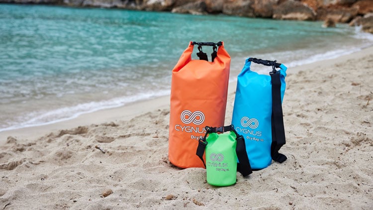 Drei Dry Bags in Orange, Blau und Grün liegen nebeneinander auf einem Sandstrand nahe dem Wasser. Alle Taschen tragen das Logo und die Aufschrift ‚CYGNUS Dry Bag‘