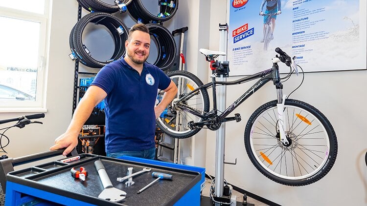Bikeservice Bikeservice. Ein junger Mann in einem blauen Shirt steht in einer Fahrradwerkstatt. Im Hintergrund sind REifen und ein Fahrrad.