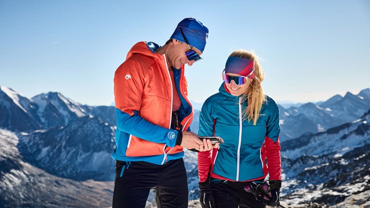 Zwei Personen in Winter-Sportkleidung stehen auf einem verschneiten Berg mit einem Gebirgszug im Hintergrund. Die linke Person trägt eine orangefarbene Jacke, eine blaue Mütze und Sonnenbrille und hält ein Gerät, auf das beide schauen. Die rechte Person trägt eivne türkisfarbene Jacke mit roten Ärmeln, ein pinkes Stirnband und ebenfalls Sonnenbrille